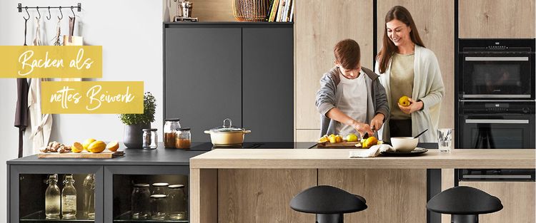 Mutter und Sohn beim gemeinsamen Kochen in moderner Küche mit Holzelementen, schwarzer Küchenzeile und der Aufschrift ‚Backen als nettes Beiswerk